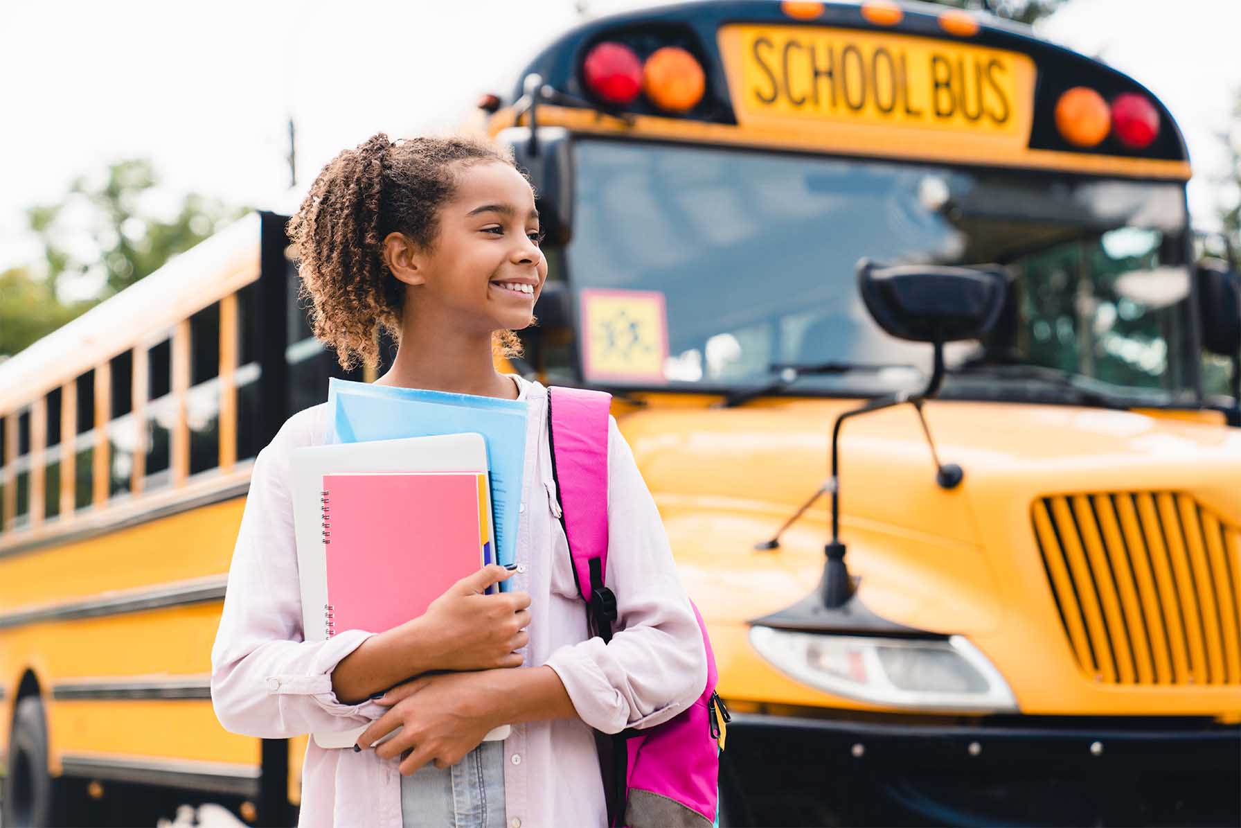 Young girl smiles holding books in front of her school bus.